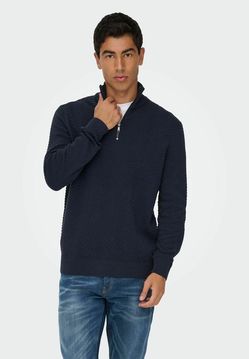 Un jeune homme portant un pull à col zippé texturé bleu marine et un jean bleu, ajustant la fermeture éclair d'une main.