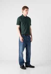 Mørkegrønn polo skjorte, blå jeans og svarte sko. Poloen har krage og tre knapper. Jeansene er rett ben med en falmet vask.