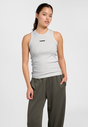 Ung kvinde iført lysegrå ærmeløs ribbet top med "hummel" tekst og løse mørkegrønne bukser, står med den ene hånd i lommen.