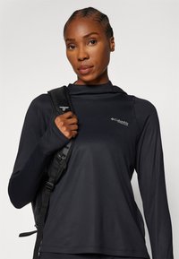 Schwarzes langärmliges Sportshirt mit strukturiertem Muster, ausgestattet mit einer Kapuze und dem Columbia-Logo. Ein schwarzer Rucksackriemen ist auf der Schulter sichtbar.
