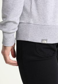 Sudadera gris con puños y dobladillo de canalé, que presenta un pequeño parche con un logo. Pantalones negros con un detalle de bolsillo. Vista en primer plano del costado.