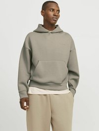 Felpa grigia con cappuccio, dal fit rilassato, in tessuto testurizzato e con tasca frontale. Indossata sopra una camicia bianca a strati e abbinata a pantaloni beige.