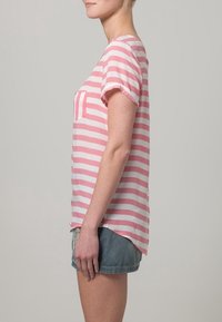 Rosa och vita randiga T-shirt med korta ärmar och en bröstficka, kombinerad med denimshorts, visad från sidan.