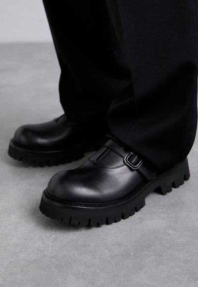 Pantofi negri din piele cu talpă groasă și detalii cu cataramă purtați cu pantaloni negri pe un podea gri.