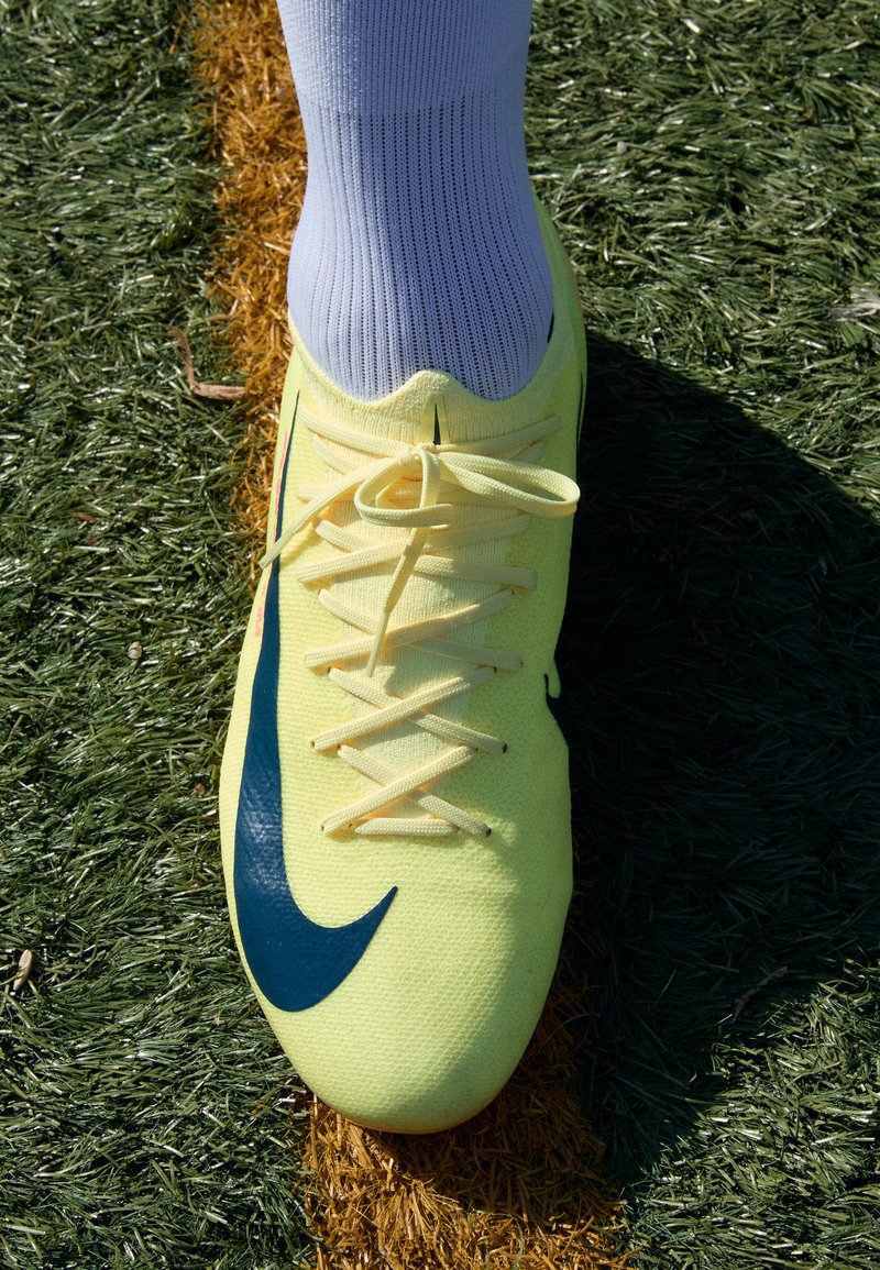 Felgele Nike voetbal schoen met een blauwe swoosh, met een gestructureerde bovenkant, platte veters, en gedragen met een witte sok op kunstgras.