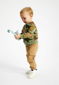 Felpa verde con stampa di dinosauro, pantaloni cargo beige e sneakers bianche; bambino che tiene in mano un giocattolo a forma di dinosauro blu.