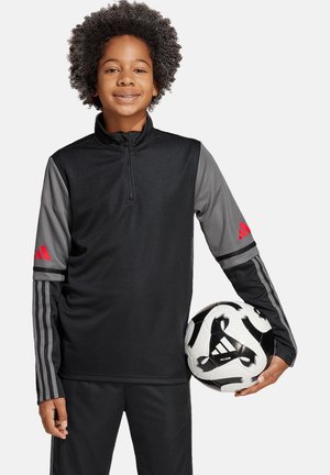 Haut de sport noir avec fermeture éclair et manches grises, portant des accents de logo rouges ; associé à un pantalon noir et tenant un ballon de soccer noir et blanc.