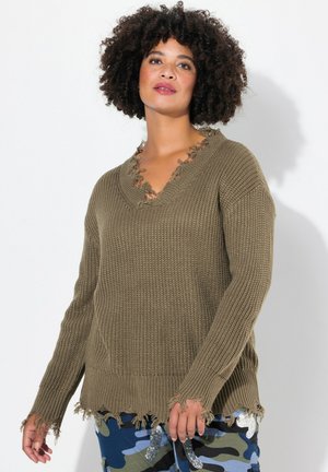 Pull en laine de couleur olive avec un col en V, aux bords effilochés et aux manches longues, associé à un pantalon en camouflage bleu et vert à motifs.