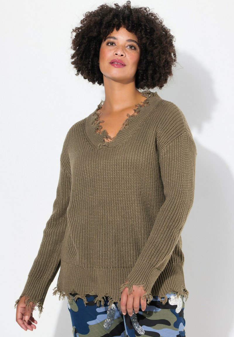 Pull en laine de couleur olive avec un col en V, aux bords effilochés et aux manches longues, associé à un pantalon en camouflage bleu et vert à motifs.
