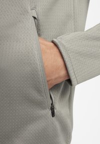 Un bolsillo con cremallera de color gris claro en una chaqueta de tela texturizada, con una mano parcialmente insertada. Presenta costuras suaves y un diseño moderno.