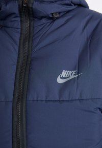 Granatowa kurtka puchowa z zamkiem błyskawicznym, z wyróżniającym się logo Nike w kolorze srebrnym. Teksturowana tkanina oraz kaptur.