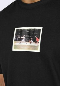 T-shirt in cotone nero con una grafica stampata rettangolare che rappresenta una scena di baseball, bordata da una cornice bianca. Collo tondo.