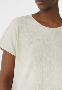 Top de manga corta color crema con una tela texturizada que presenta sutiles patrones en forma de ondas y un escote redondo. Diseño sencillo y de corte relajado.