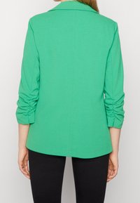 Blazer verde con un diseño ajustado, mangas tres cuartos fruncidas y un cuello clásico. Textura suave, forma entallada y parte trasera alargada.