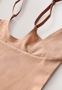 Bezrukávové ľahko hnedé ľanové šaty s nastaviteľnými tenkými ramienkami a výstrihom do V, rozložené na bielom povrchu.