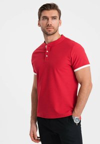 Červené polo triko s texturovaným povrchem, krátké rukávy s bílými akcenty a tříknoflíkovou légou. Nosené s černými kalhotami.
