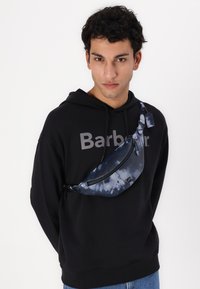 Borsetă tip fanny pack cu un model tie-dye albastru, cu o textură netedă, închidere cu fermoar și o curea neagră ajustabilă, purtată peste un hanorac negru cu litere gri.
