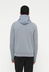 Grå mønstrete hettegenser med en glatt, lysere grå hette. Har et rutenettdesign, ribbede mansjetter og en nederkant. Bakutsyn, brukt av en person.