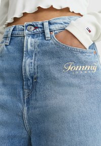 Jeans de denim azul claro con cintura alta, caracterizados por un botón plateado, logo bordado "Tommy Jeans" y diseño clásico de bolsillos.