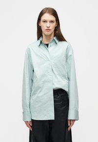 Mujer joven con cabello largo y castaño, vestida con una camisa azul claro de botones oversize y pantalones negros, posando frente a un fondo blanco liso.
