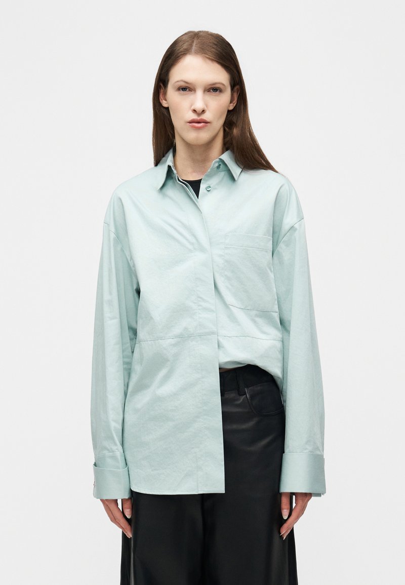 Νεαρή γυναίκα με μακριά καστανά μα λλιά που φοράει ένα ανοιχτό γαλάζιο, φαρδύ πουκάμισο με κουμπιά και μαύρο παντελόνι, στέκεται μπροστά σε ένα απλό λευκό φόντο.