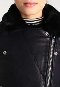 Černá kožená bunda s umělým kožešinovým límcem, stříbrnými zipy a druky. Vnitřní vrstva má texturovaný, pruhovaný design.
