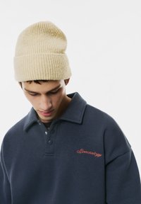 Mørkeblå sweatshirt med foldet krave, prydet med røde broderede bogstaver. En lys beige ribbet strikhue sidder ovenpå modellens hoved.