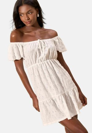 Femeie purtând o rochie albă cu broderie, cu umerii goi, mâneci scurte și tiv cu volane, privind în lateral cu un zâmbet subtil.