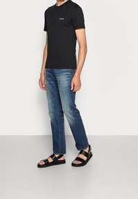 Svart t-shirt med kort ärm, blå denim jeans med blekning och svarta sandaler med remmar. Platt sula, justerbara ankelremmar och öppen tå.