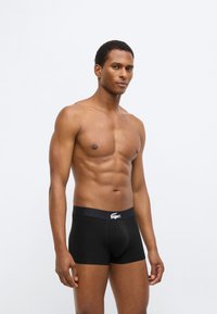 Μαύρα μποξεράκια με λωρίδα μέσης με λογότυπο. Το ύφασμα φαίνεται λείο και εφαρμοστό, παρουσιάζοντας έναν μινιμαλιστικό σχεδιασμό.