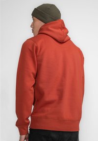 Felpa rossa con tessuto strutturato, dotata di tasca a marsupio e cappuccio con coulisse. Sulla testa è indossato un berretto verde.