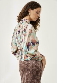 Blusa floral en colores pastel con mangas abullonadas y cintura ajustada; tejido suave con acabado brillante. Combinada con una falda marrón texturizada.