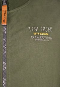 Oliwkowa bluzka z zamkiem błyskawicznym, wyszytym napisem "TOP GUN" oraz skórzaną metką. Miękka tkanina o gładkiej fakturze.