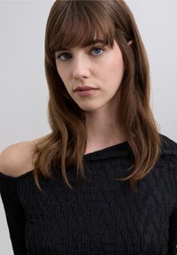 Musta olkapäitä paljastava pusero, jossa on aaltoilevaa kuvioivaa tekstuurikangasta. Kapea malli ja pitkät hihat, korostettu hienovaraisella kiillolla.