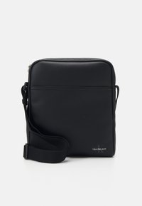 Bolso cruzado sintético negro con textura suave, cierre de cremallera, correa ajustable y detalle de logo grabado de Calvin Klein.