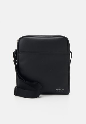 Bolso cruzado sintético negro con textura suave, cierre de cremallera, correa ajustable y detalle de logo grabado de Calvin Klein.