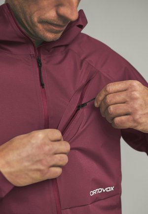 Mann trägt eine kastanienbraune Ortovox-Jacke und öffnet die Brusttasche mit einem Reißverschluss-Zuglaschen vor einfarbigem Hintergrund.