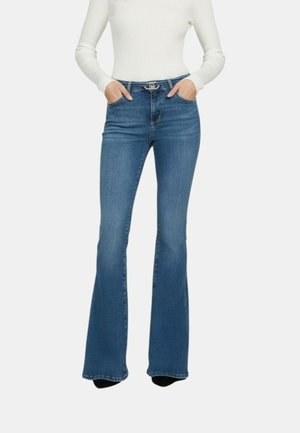 Persona che indossa jeans blu a vita alta con gamba svasata, tasche frontali e cintura nera, abbinati a una maglia bianca a coste con maniche lunghe, mani nelle tasche.