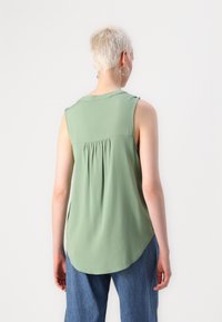 Ermeløs grønn topp laget av glatt stoff, med rund halslinning og rynkede detaljer bak. Kombinert med blå jeans.