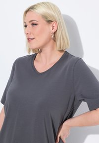 Blonde vrouw met kort haar, die zilveren creolen en een grijze V-hals T-shirt draagt, kijkt naar links met haar hand op haar heup tegen een witte achtergrond.