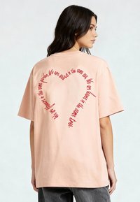 Šviesiai rožinė per didelė medvilninė marškinė, ant nugaros su raudonu kursyvu parašyta tekstas, sudaranti širdies formą. Trumpi rankoviai ir apvalus kaklas.