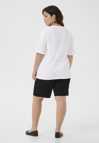 Chemise blanche à manches courtes avec accents texturés sur les épaules, associée à un short noir et des ballerines noires. Design simple et coupe décontractée.