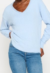 Pull bleu clair duveteux avec un col en V, des manches longues et un ourlet légèrement arrondi. Porté avec un jean bleu ajusté. Texture douce.