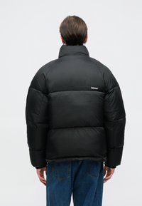 Μαύρο μπουφάν με ψηλό γιακά, με διάταξη που έχει κατασκευή από καπιτονέ υλικό και ελαστικούς καρπούς. Στην πλάτη υπάρχει ένα μικρό λογότυπο σε λευκό χρώμα.