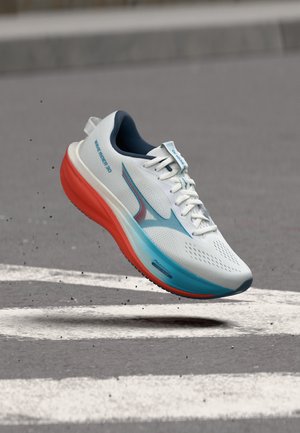 Chaussure de course blanche et bleue avec semelle rouge en l'air au-dessus de l'asphalte avec des marquages routiers blancs.