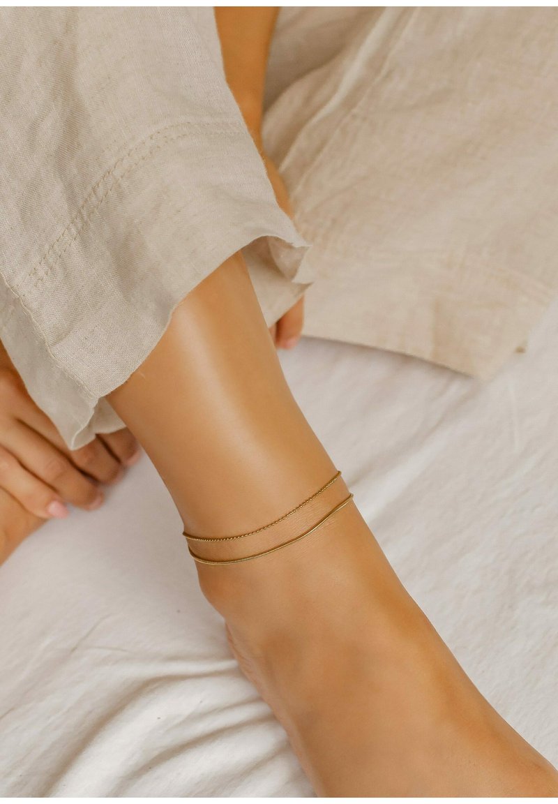 Pulsera de tobillo de oro con dos delicadas cadenas que envuelven un tobillo liso, sobre un tejido de lino de color claro.