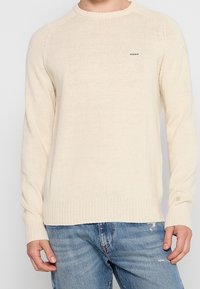 Cremfarbener Strickpullover mit rundem Ausschnitt, gerippten Bündchen und Saum sowie einem kleinen Logopatch auf der Brust. Getragen über hellblauen Jeans.