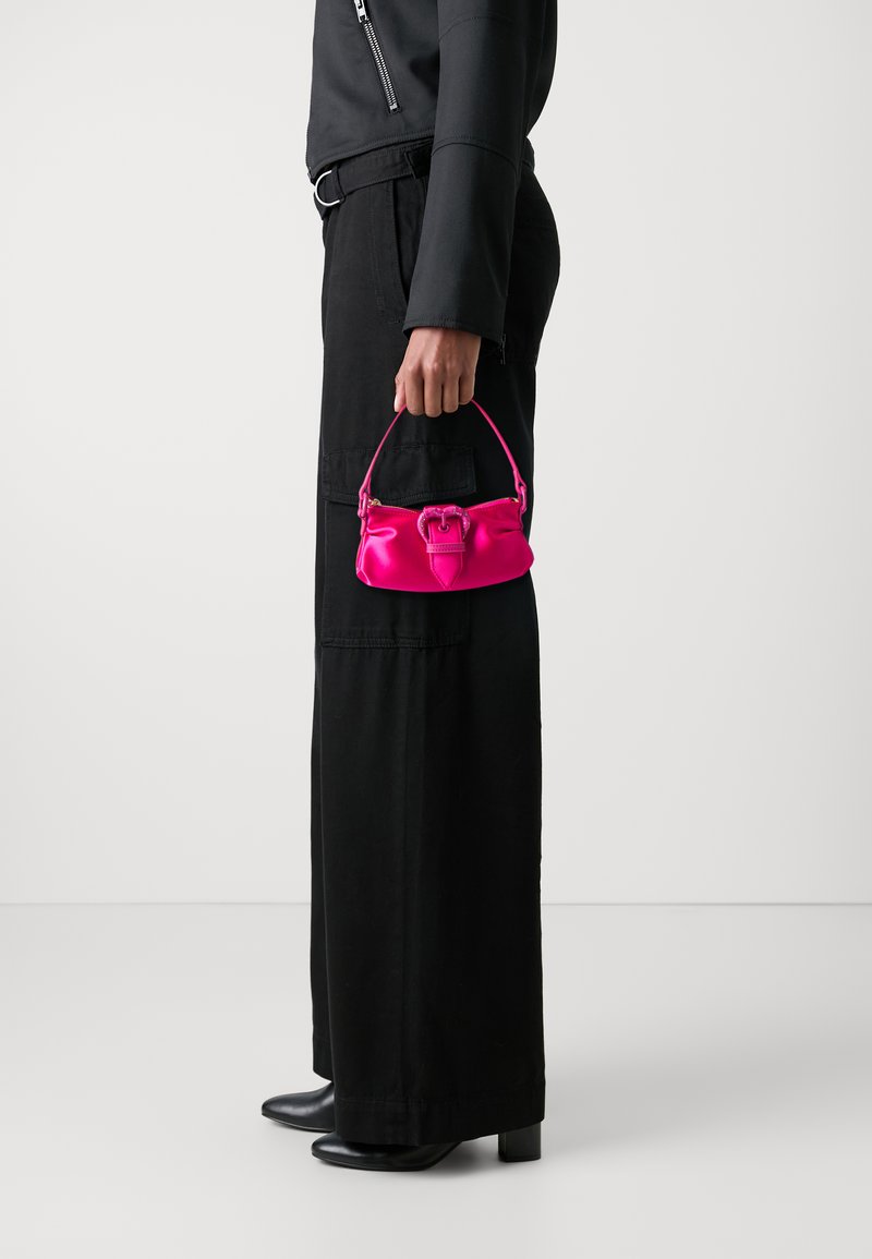 Ζεστό ροζ σατέν τσάντα χειρός με δομημένο σχήμα και πάνω χερούλι. Περιέχει λεπτομέρεια κλεισίματος σε στυλ φιόγκου και κρατιέται από ένα άτομο ντυμένο στα μαύρα.