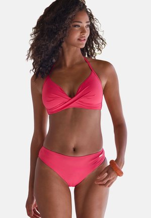 Frau mit lockigem Haar, die einen pinkfarbenen Neckholder-Bikini und ein orangefarbenes Armband trägt und nach rechts schaut, vor weißem Hintergrund.