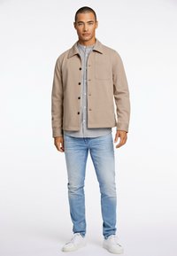 Beige Jacke mit Kragen, Knopfleiste und einer Brusttasche, über einem gestreiften Hemd getragen, kombiniert mit hellblauen Jeans und weißen Sneakern.